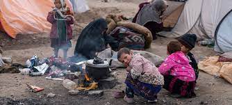 A child is crouching next to a kettle boiling on a open pit wood-fire. The child is looking over their shoulder to the person taking the picture. There are personal belongings scattered on the floor around the child.