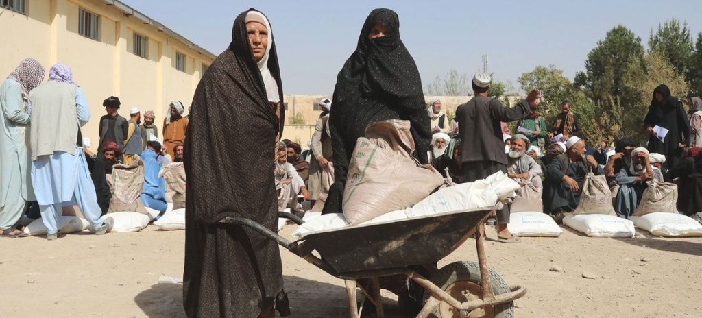 Two people clad in women's garb (long black dresses covering the head). One is holding a wheelbarrow filled with white bags. Some other people are visible in the background. The picture was taken outside on a sunny day.