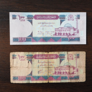 Two Afghani banknotes are placed side by side on a flat surface. The upper one looks brand new, the lower one is worn out and partly torn.