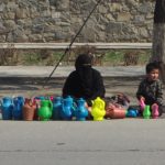 Mother and child sitting on the floor beside a road. In front of them there are colourful plastic jars, teapots, and cups. It is sunny and the earth is completely parched brown.