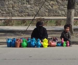 Mother and child sitting on the floor beside a road. In front of them there are colourful plastic jars, teapots, and cups. It is sunny and the earth is completely parched brown.