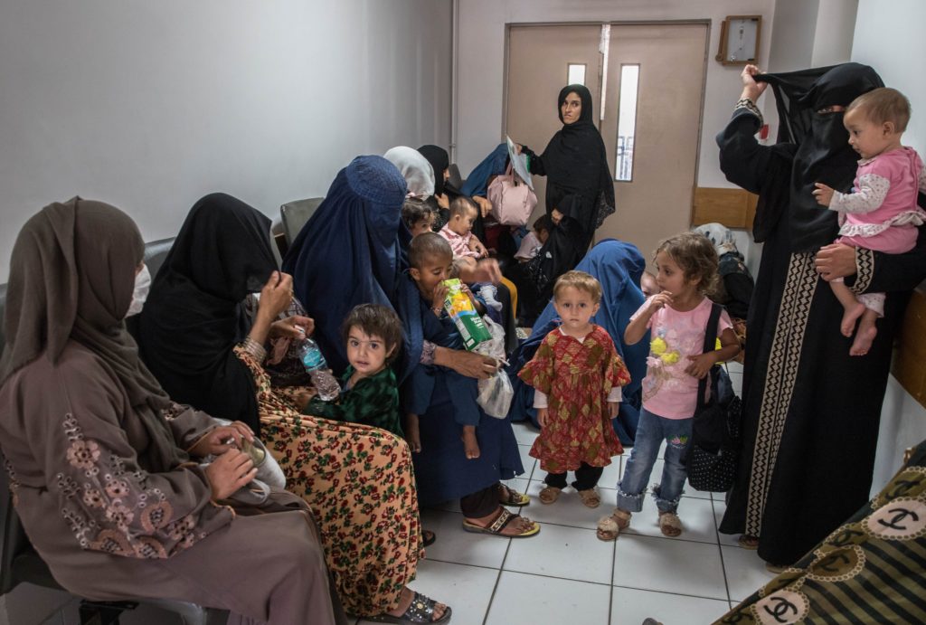 Women, children and babies crowded into a corridor. 6 women are sitting on chairs lining the left corridor wall and two women are standing.