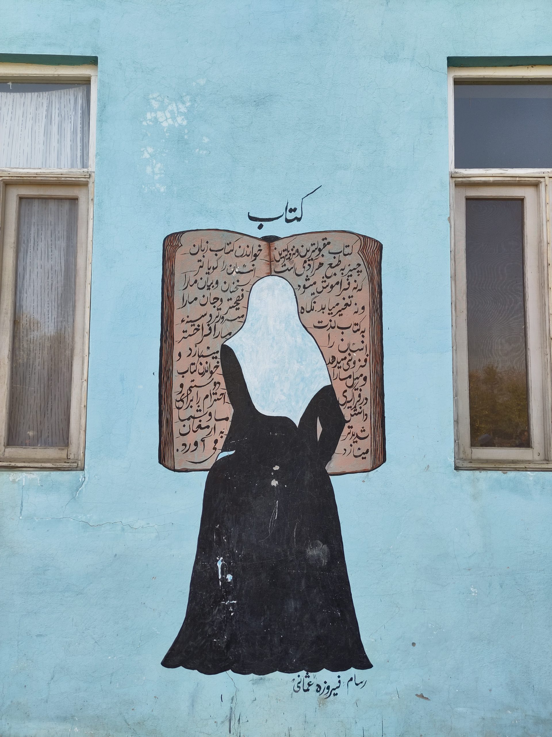 A wall painting of the back of a student wearing a woman's uniform. The student is painted standing in front of huge open book. Above the painting the word "Book" is written in Dari. There are two windows visible on each side of the wall painting.