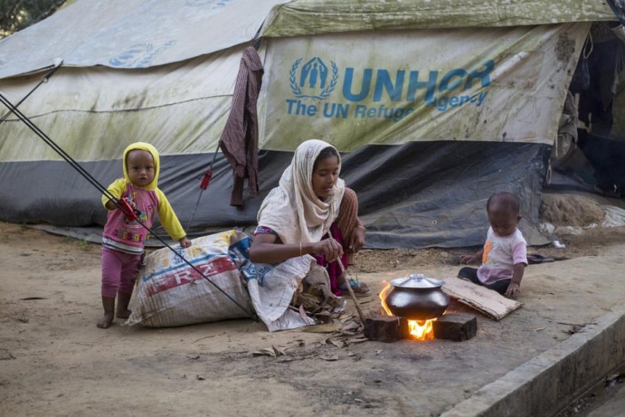 A woman sits outside, next to a fire on the ground. A kettle sits on the fire. Two children stand around and look at the camera. In the background there is a large tent with the UNHCR logo on it.