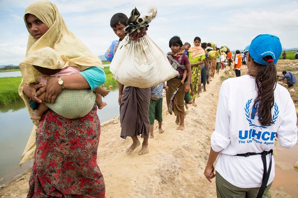 Women and children walking on a dirt path in single file, carrying big bags made of cloth in their arms. One person stands next to the queue watching the people pass. This person is wearing a UNHCR T-shirt and a blue cap.