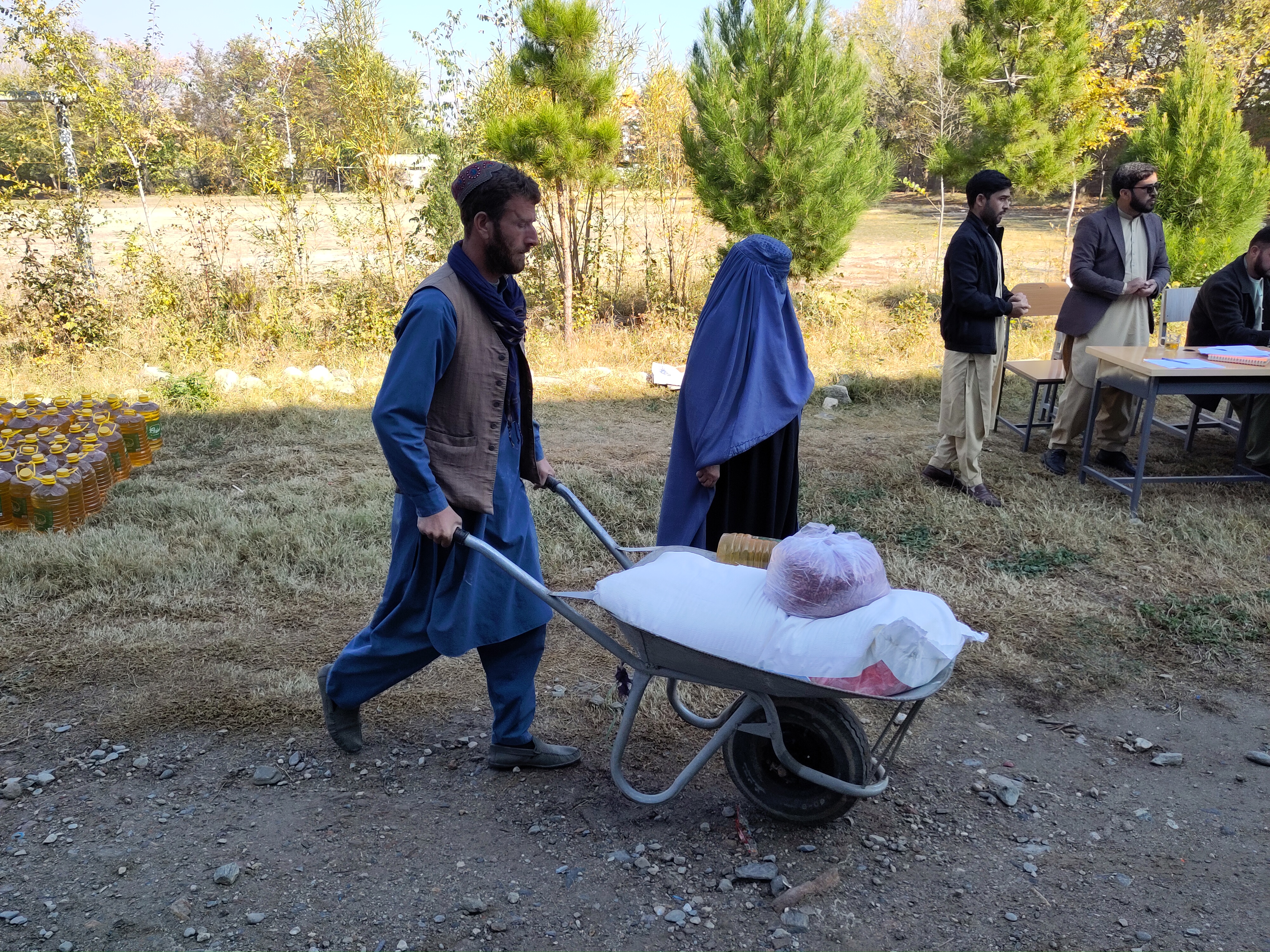 A man holds a wheelbarrow with bags of food in it. A woman walks by his side. In the background there is a table with sheets of paper and men standing and waiting.