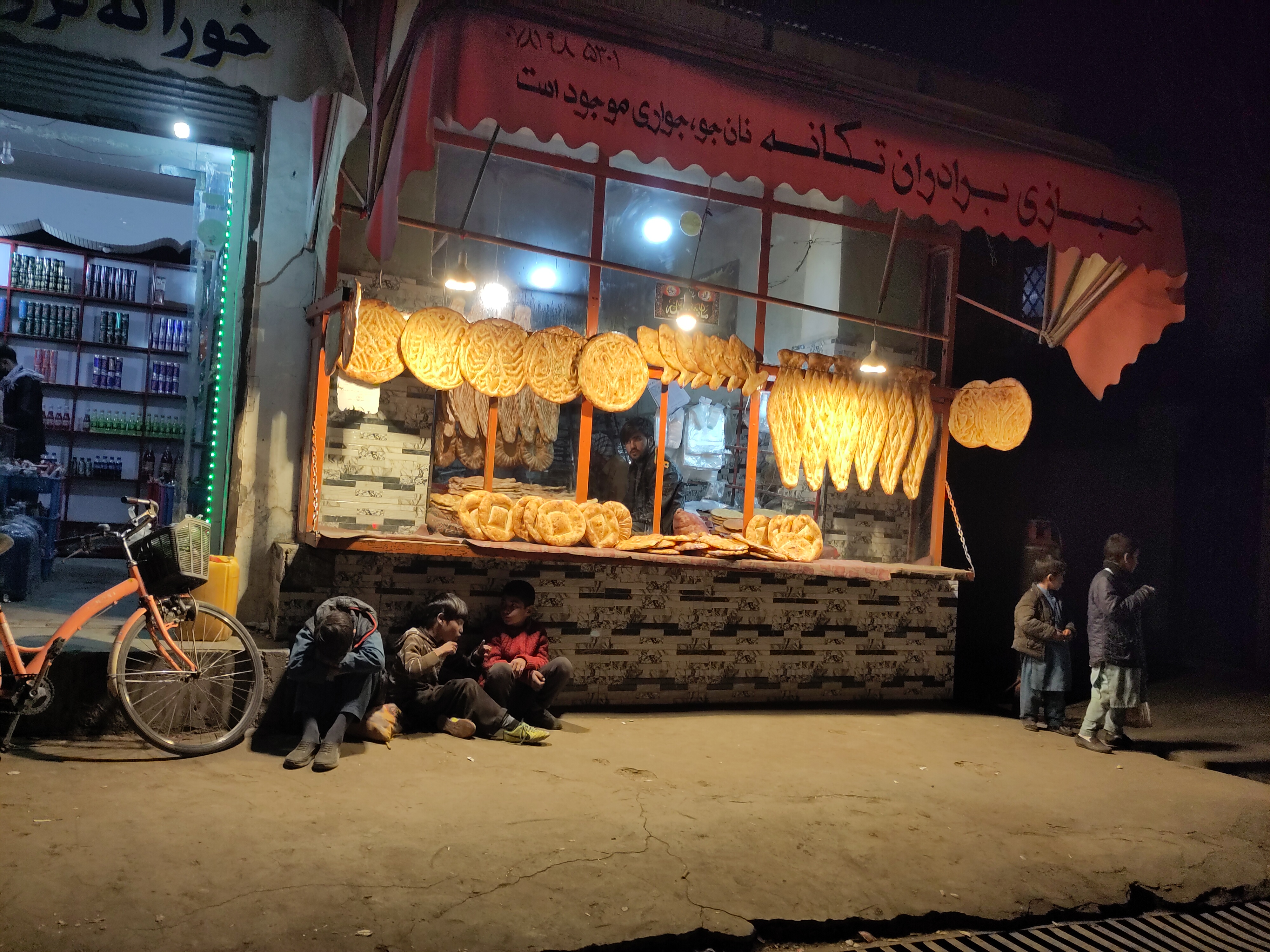 The store front of a bakery is lined with bread hanging, lit by a few lightbulbs in the night. Some children are sitting on the street floor and looking out on onto the street.