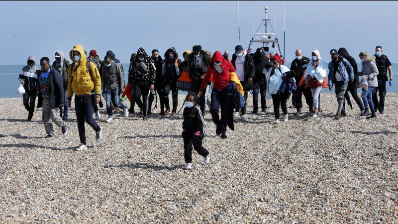 A group of 30 or more people walk head down on a beach. A few children wearing facemasks are at the front of the group. Behind the group there is at least one uniformed British police officer and the silhouette of a boat.