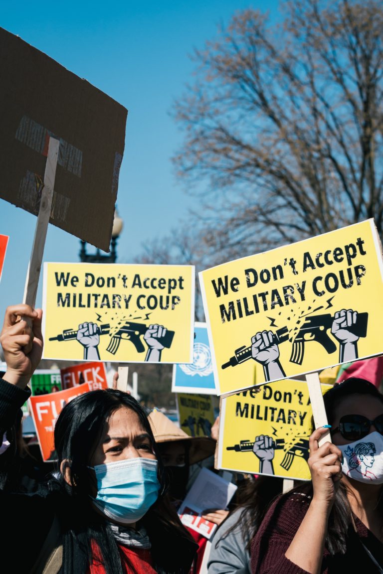 A close-up on the faces (with Covid masks) of a crowd of people holding up yellow signs with "We Don't Accept Military Coup" written in black letters.