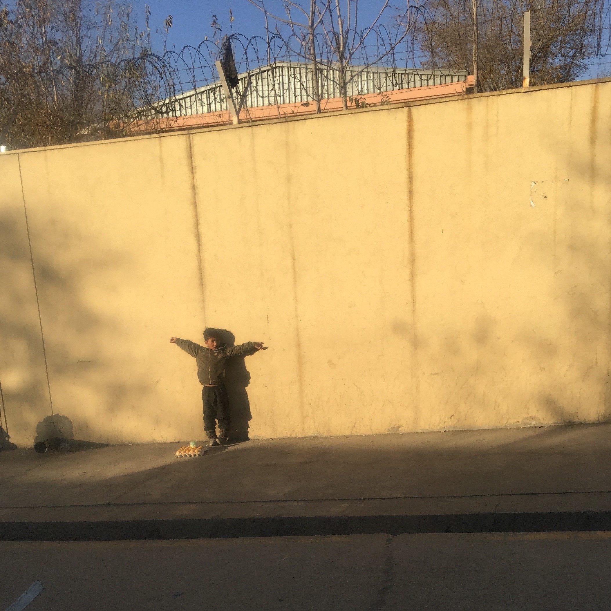 Boy standing in the street with arms raised in a T, against a wall.